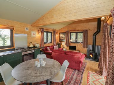 A living area with a round dining table and chairs a kitchen with green cabinets and a stove and a sitting area with red sofas a wood burning stove and windows with curtains at Willow in Dunchurch