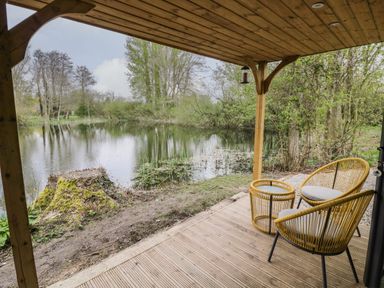 A covered wooden patio with two chairs and a table overlooking a pond surrounded by trees at Dragonfly in Dunchurch