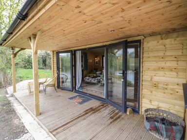 A wooden porch with two yellow chairs and a table outside a cabin with glass doors at Dragonfly in Dunchurch