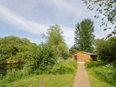 A wooden cabin at the end of a gravel path surrounded by trees and grass at Dragonfly in Dunchurch