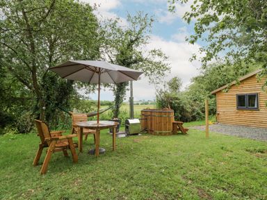 An outdoor garden area with wooden chairs around a table with an umbrella a wooden hot tub a hammock and a wooden building at Dragonfly in Dunchurch