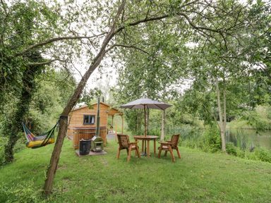 An outdoor area with wooden chairs around a table and umbrella a hammock and a small wooden cabin near a body of water at Dragonfly in Dunchurch