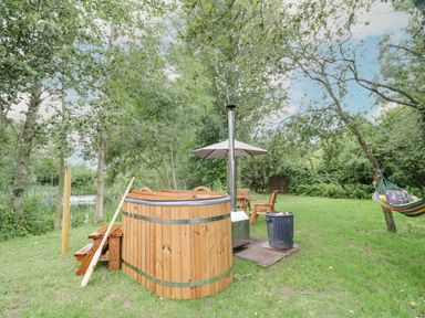 An outdoor area with a wooden hot tub, a small wooden table and chairs under an umbrella, and a hammock tied between trees at Dragonfly in Dunchurch