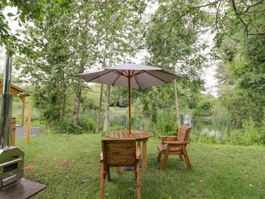 A wooden table with two wooden chairs and a large umbrella on grass near trees and a pond at Dragonfly in Dunchurch