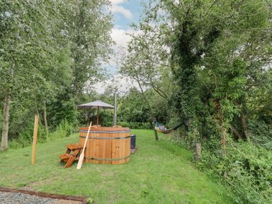 An outdoor area with a wooden hot tub umbrella and hammock among trees and grass at Dragonfly in Dunchurch