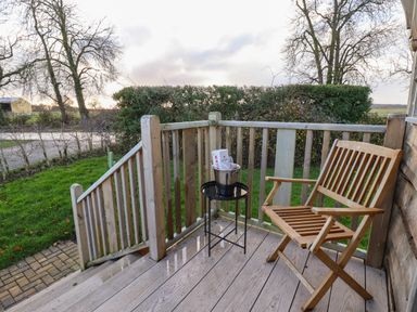 A wooden chair and small table with a metal bucket on a wooden deck outside a house with trees and grass around at Arts and Crafts in Bolton Percy near Tadcaster