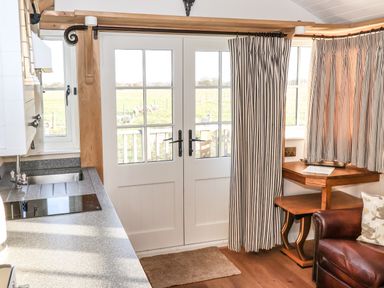 A kitchen area with a sink and stove next to glass doors with curtains and a corner wooden table by a leather armchair at The Egyptologist in Bolton Percy near Tadcaster