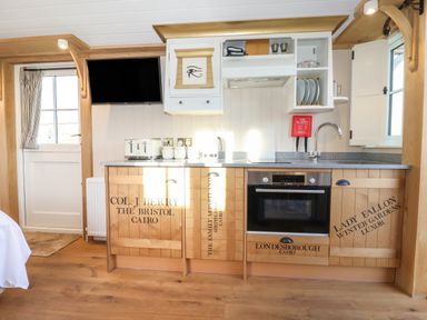 A kitchen area with wooden cabinets, an oven, a sink, a countertop with a toaster and kettle, a wall-mounted TV, and a cabinet with plates at The Egyptologist Bolton Percy near Tadcaster