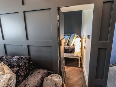 A room with a dark textured couch and an open door leading to a small bedroom with a bed and pillows at Whisk Away Cottage Greystoke near Troutbeck Near Penrith