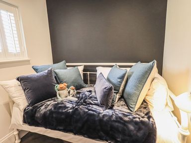 A small bed with multiple blue and gray pillows and two plush toys in a room with a dark gray wall and window at Whisk Away Cottage Greystoke near Troutbeck Near Penrith