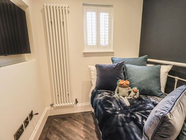 A bedroom corner with a bed covered in cushions and two stuffed frogs near a window and a radiator at Whisk Away Cottage in Greystoke near Troutbeck Near Penrith