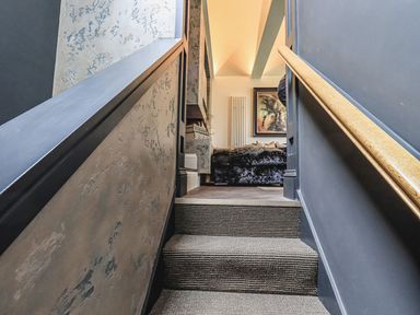 A carpeted staircase with dark walls and a wooden handrail leading to a room with a sofa and a framed painting at Whisk Away Cottage in Greystoke near Troutbeck Near Penrith