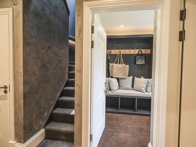 A hallway with carpeted stairs on the left and a room on the right with a seating bench with cushions and hooks holding bags