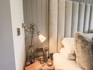 A bedside table with books vases and a lamp next to a bed with a cushioned headboard at Whisk Away Cottage in Greystoke near Troutbeck Near Penrith