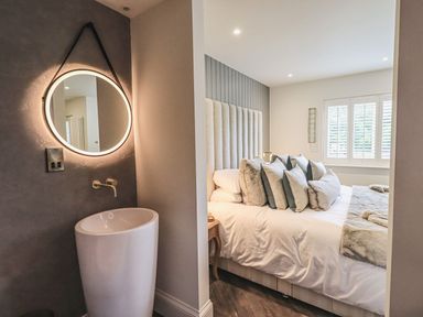 A bedroom with a bed and pillows next to a space with a tall sink and round illuminated mirror at Whisk Away Cottage in Greystoke near Troutbeck Near Penrith