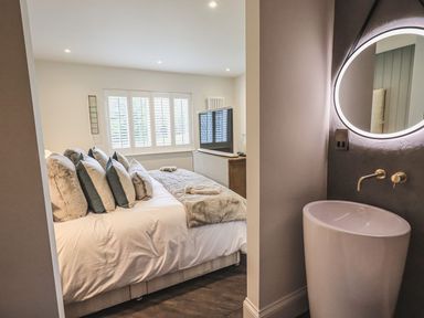 A bedroom with a bed multiple pillows a television and a bathroom sink with a round backlit mirror at Whisk Away Cottage in Greystoke near Troutbeck Near Penrith