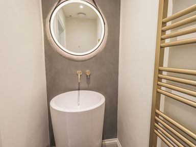 A bathroom with a round illuminated mirror above a modern pedestal sink and a gold towel radiator at Whisk Away Cottage in Greystoke near Troutbeck Near Penrith