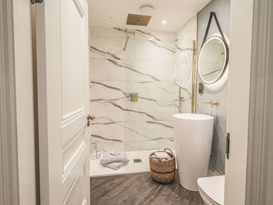 A bathroom with a walk-in shower towels on the floor a wicker basket a tall white sink and a round mirror at Whisk Away Cottage in Greystoke near Troutbeck Near Penrith