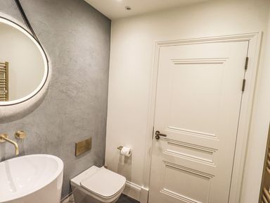 A bathroom with a round illuminated mirror a sink a wall-mounted faucet a toilet a toilet paper holder and a white door at Whisk Away Cottage in Greystoke near Troutbeck Near Penrith