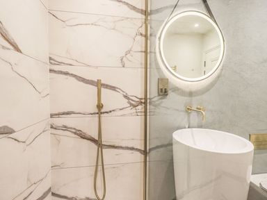 A bathroom with marble walls a round illuminated mirror a white sink and a gold shower head at Whisk Away Cottage in Greystoke near Troutbeck Near Penrith