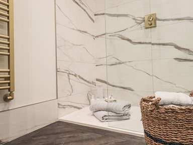 A shower area with marble-patterned tiles a glass shower screen a stack of folded towels and toiletries and a wicker basket with towels at Whisk Away Cottage in Greystoke near Troutbeck Near Penrith