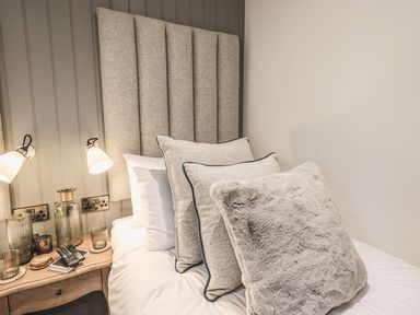 A bed with multiple pillows and a wooden bedside table with lamps and glassware at Whisk Away Cottage in Greystoke near Troutbeck Near Penrith