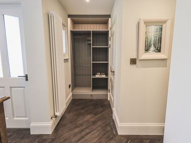 A hallway with a built-in closet shelves hangers a white door a radiator and a framed forest painting at Whisk Away Cottage Greystoke near Troutbeck Near Penrith
