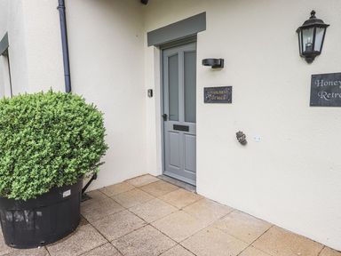 A grey entrance door with signs on cream exterior wall and a large potted bush outside at Whisk Away Cottage Greystoke near Troutbeck Near Penrith
