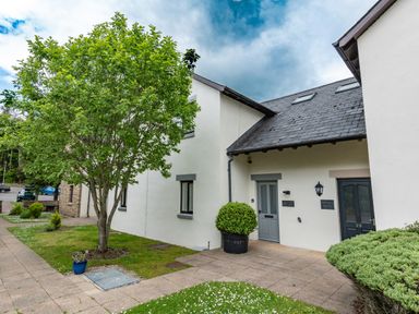 Exterior view of white buildings with grey doors and windows a tree and bushes on a paved walkway at Whisk Away Cottage in Greystoke near Troutbeck Near Penrith