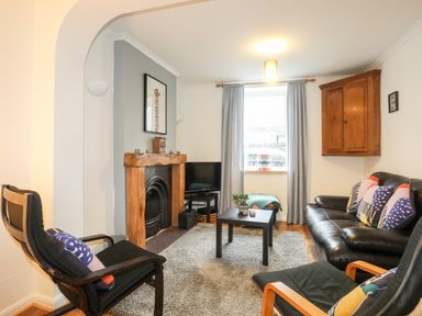 A living room with a black leather sofa and chairs a wooden fireplace a small black coffee table and a window with grey curtains at 5 Thomas Street in Llanberis