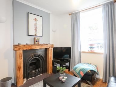 A living room with a fireplace wooden mantel a black TV on a stand a window with gray curtains an ottoman with a colorful blanket and a black coffee table with plants and candles at 5 Thomas Street in Llanberis
