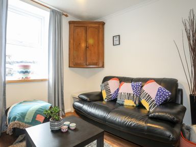 A living room with a black leather sofa with three colorful pillows a small black coffee table with a plant and candles a wooden corner cabinet and a window with grey curtains at 5 Thomas Street in Llanberis