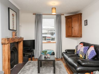 A living room with a black leather sofa pillows a wooden fireplace a black coffee table a television on a stand gray curtains and a wall cabinet at 5 Thomas Street in Llanberis