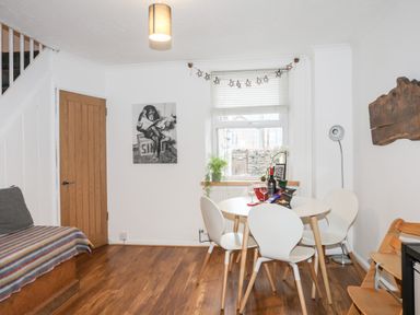 A dining area with a round table and four chairs near a window with blinds a floor lamp a wooden door and a staircase at 5 Thomas Street in Llanberis