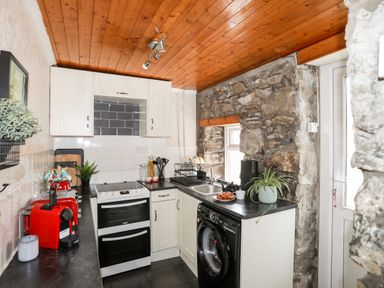 A kitchen with stone walls a wooden ceiling white cabinets and black appliances at 5 Thomas Street in Llanberis