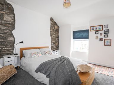 A bedroom with a bed between exposed stone walls a window with a dark blind and picture frames on the wall at 5 Thomas Street in Llanberis