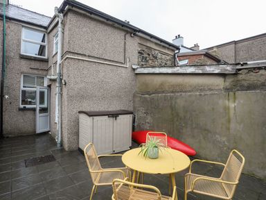 An outdoor patio area with yellow metal table and chairs a potted plant on the table a red kayak leaned against a concrete wall and a plastic storage box by a house at 5 Thomas Street in Llanberis