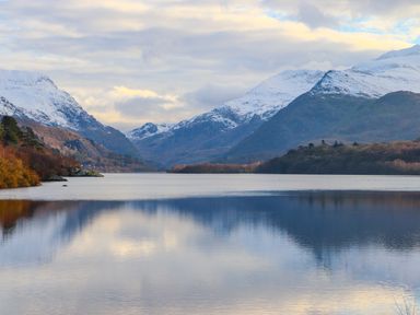 A lake with snow capped mountains and trees reflected in the water at 5 Thomas Street in Llanberis
