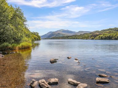 A lake with rocks in the water trees along the shore and hills in the background at 5 Thomas Street in Llanberis