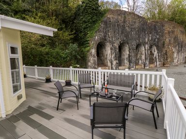 An outdoor deck with chairs a table with a bottle and glasses overlooking a stone wall and greenery at Seacroft in Stepaside near Kilgetty