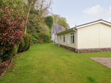 A side view of a single-story house with yellow siding and a stone foundation next to a green lawn with bushes and trees at Seacroft in Stepaside near Kilgetty