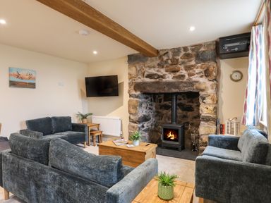 A living room with grey sofas a stone fireplace with a wood burner a wooden coffee table and a wall mounted television at Pennant Igyn in Mynytho near Abersoch