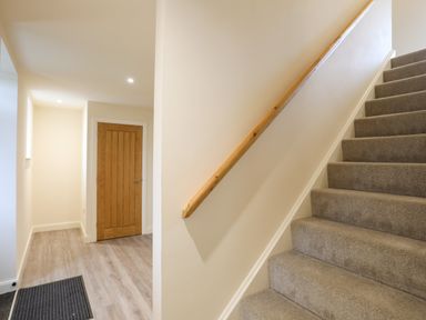 A hallway with wood flooring and a wooden door next to carpeted stairs with a wooden handrail at Pennant Igyn in Mynytho near Abersoch