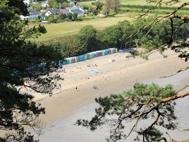 A sandy beach with colorful beach huts along the edge people on the beach and trees framing the view at Pennant Igyn in Mynytho near Abersoch