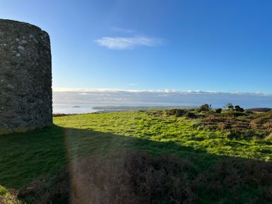 A stone structure on grassy land with a distant view of the sea and sky at Pennant Igyn in Mynytho near Abersoch