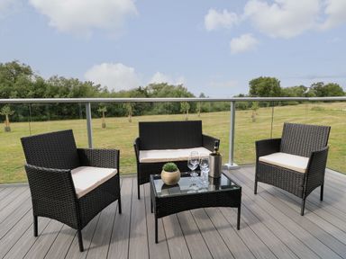 An outdoor seating area with black wicker chairs and a table with glasses and a bottle on a wooden deck overlooking a grassy field at Holly Lodge in Winthorpe near Newark-On-Trent