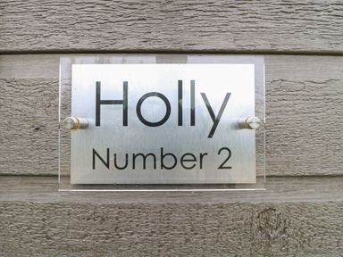 A metal and acrylic house sign on a textured wooden wall with the text Holly Number 2