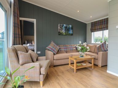 A living room with a beige sofa and armchair with plaid cushions a wooden coffee table with a flower vase and a framed forest picture on the wall at Cedar Lodge in Newark-On-Trent