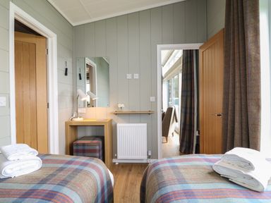 A bedroom with two single beds with plaid blankets and folded towels a wooden desk with a lamp and a stool a door leading to another room with chairs at Cedar Lodge in Newark-On-Trent
