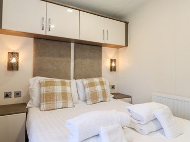 A bedroom with a double bed two checkered pillows folded towels bedside tables wall lamps and overhead cabinets at Orion in Felmoor Park